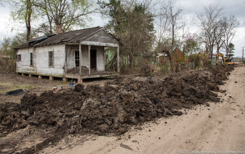 Construction equipment tears up a dirt road to replace water pipes next to a ramshackle house.