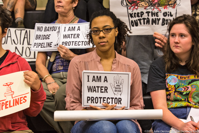 Women sit in the stands at the permit hearing, holding signs opposing the Bayou Bridge pipeline