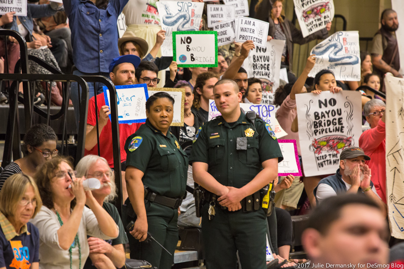 Two law enforcement officers stand among the stands of pipeline opponents holding signs at a permit hearing