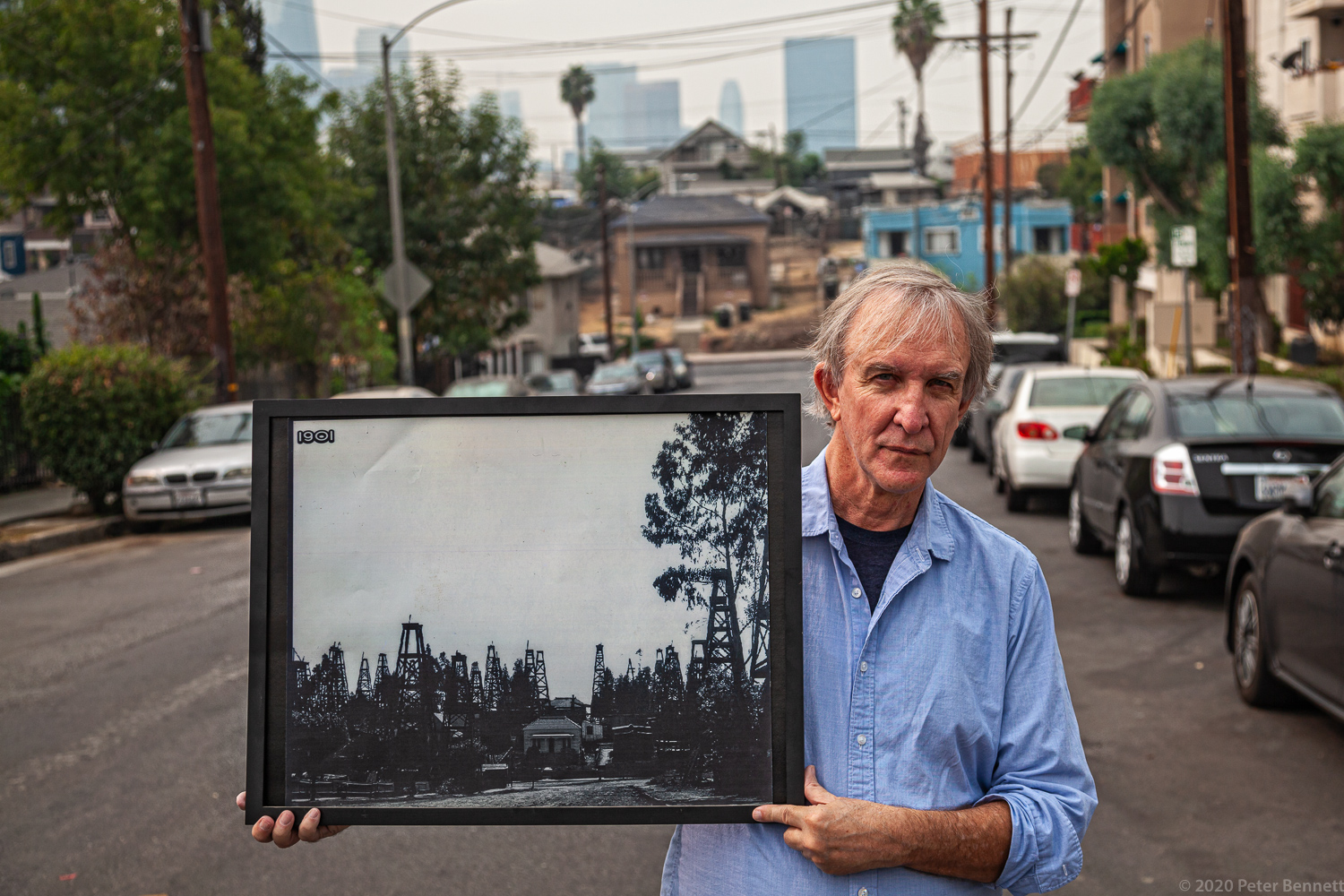 Resident, Thomas Gustafson holds a photo from 1901 in front of an original home on Edgeware Road and Court Street, once surrounded by oil derricks.