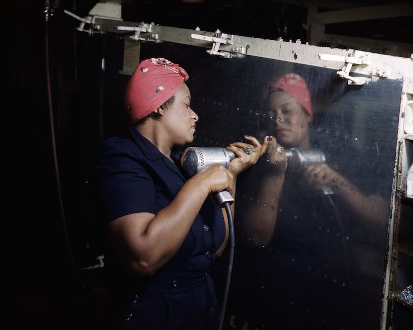 A woman dressed in work coveralls and a red bandanna on her head operates a hand drill on a war plane during World War II